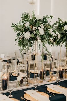 an elegant table setting with tall vases filled with white flowers and greenery on top