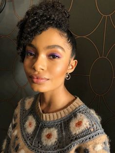 a woman with short hair and purple eyeshadow is posing for a photo in front of a wall