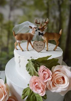 two deer figurines on top of a white cake with pink roses and greenery