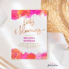 a pink and gold wedding card on top of a white surface with straw hat next to it