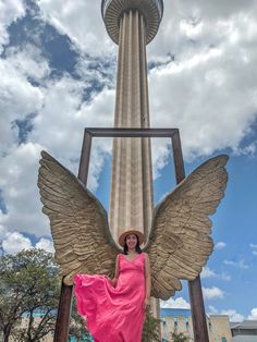 a woman in a pink dress and hat standing next to an angel statue with the space needle behind her