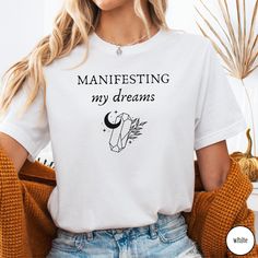 Embrace the power of manifestation with our 'Manifesting My Dreams' Crystal Shirt. Designed with witchy, mystical vibes, this tee blends the energy of crystals with the Law of Attraction. Whether you're manifesting your desires or drawn to spiritual practices, this shirt offers both style and inspiration. Perfect for dreamers, spiritual seekers, or crystal lovers, it also makes a thoughtful gift. Would you prefer this design on a Sweatshirt? Have a look at our other listings here:  https://raidh Spiritual Tee Shirts, Cheap Spiritual Graphic Print T-shirt, Crystal Shirt, Power Of Manifestation, Boho Frame, Spiritual Manifestation, The Law Of Attraction, Spiritual Practices, Jersey Shorts