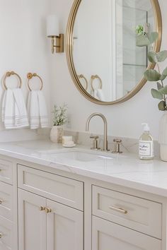 a white bathroom with gold accents and marble counter tops, an oval mirror above the sink