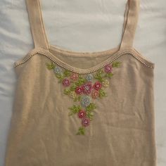 a tan top with pink and green flowers embroidered on the front, sitting on a white sheet