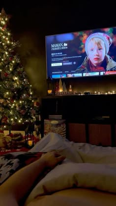 a person laying in bed with a christmas tree next to it and a large screen on the wall