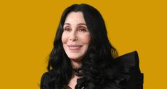 a woman with long black hair wearing a black dress and smiling at the camera while standing in front of a yellow background