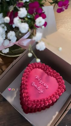 a pink heart shaped cake sitting in a box on top of a table next to flowers