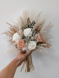 a hand holding a bouquet of flowers on top of a white wall with dry grass