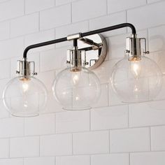 three clear glass globe lights hanging from a metal fixture on a white tiled bathroom wall