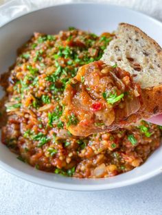 a white bowl filled with meat and vegetables on top of a wooden spoon next to bread