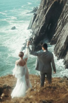 a bride and groom walking along the cliff by the ocean