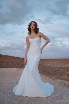 a woman standing on top of a dirt field wearing a white dress and holding her hands on her hips