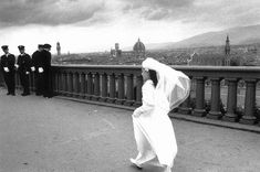 black and white photograph of woman in wedding dress walking on bridge with men standing behind her