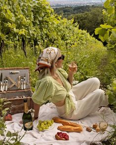 a woman sitting on a blanket eating food and drinking wine in front of some trees