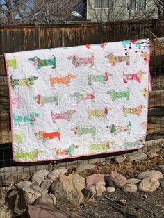 a quilt hanging on a fence in front of some rocks and trees with a house in the background