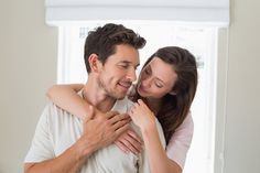 a man and woman embracing each other in front of a white window with blinds on the windowsill