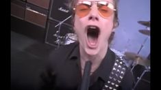 a man wearing sunglasses and singing into a microphone in front of a wall with musical instruments