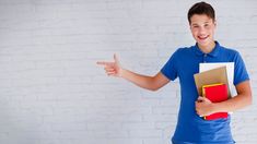 a young man is holding books and pointing to the side with his hand while standing in front of a white brick wall