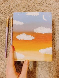 a hand holding a pen and drawing clouds on a piece of paper with pastel pencils next to it