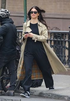 a woman in black pants and trench coat walking down the street next to a man