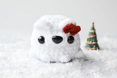 a small white stuffed animal sitting in the snow next to a tiny christmas tree with black eyes