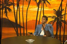 a man sitting at a table in front of palm trees with an orange sunset behind him