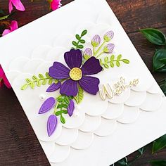 a white card with purple flowers and green leaves on it, next to some pink flowers