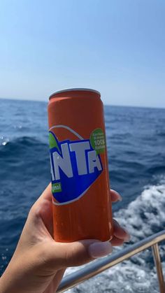 a person holding up a can of orange soda in front of the ocean on a boat