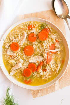 a bowl of soup with carrots, chicken and rice on a white surface next to a spoon