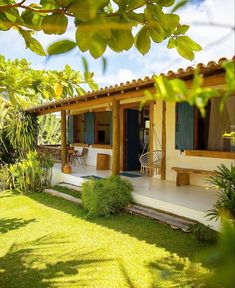 a small house with blue shutters and green plants on the front lawn, surrounded by greenery