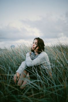 a woman sitting in tall grass on top of a field with her arms folded over her chest