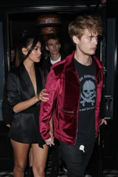 a man in a red velvet jacket and black shorts is walking towards the camera with a woman