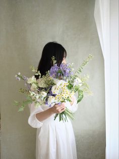 a woman holding a bouquet of white and purple flowers in her hands while standing next to a window