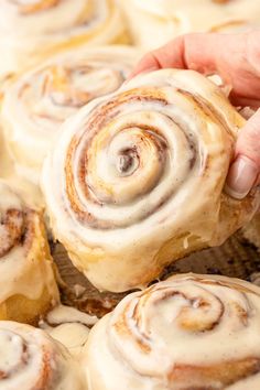 a person holding a cinnamon roll in front of some rolls on a baking sheet with icing