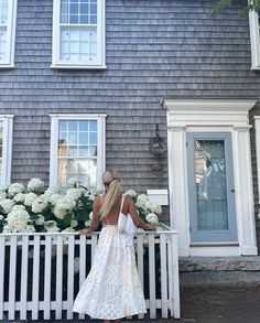 a woman in a white dress standing on a porch next to a house with flowers