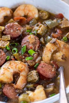 a white bowl filled with shrimp and sausage stew next to a spoon on a wooden table