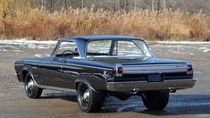 an old black car parked in a parking lot with snow on the ground behind it