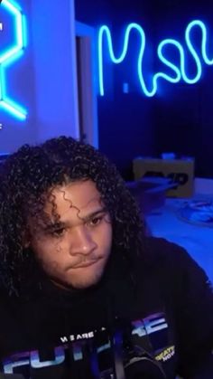 a man with long curly hair sitting in front of a blue neon sign that says masu