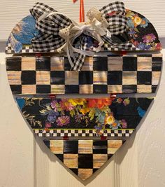 a wooden heart hanging on the wall with flowers and checkered ribbon tied to it