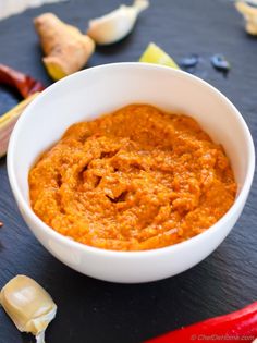 a white bowl filled with carrot hummus next to garlic and red peppers on a table