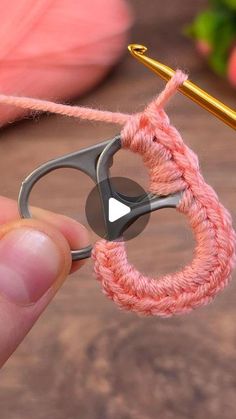 a person holding a pair of scissors in front of a piece of pink yarn with a crochet hook