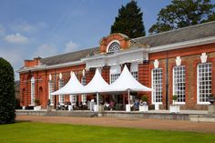 an old brick building with white tents on the front lawn and people sitting at tables under umbrellas