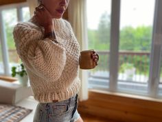 a woman holding a coffee cup in her right hand and looking at the camera while standing next to a window