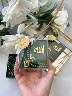 a hand holding a green box with gold lettering on it and flowers in the background