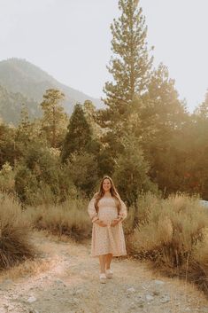 a woman standing on a dirt road in front of some tall trees and grass with mountains in the background