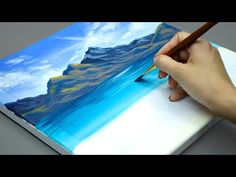 a person is holding a pencil and drawing on a piece of paper with blue mountains in the background