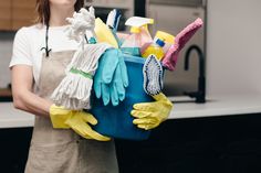 a woman in an apron and rubber gloves holding a bucket full of cleaning supplies on the kitchen counter