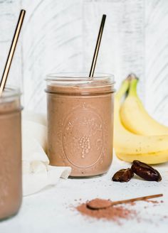 two jars filled with chocolate smoothie next to bananas and cinnamon on a white table