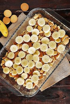 a glass dish filled with banana slices and nuts on top of a wooden table next to two bananas