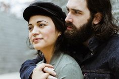 a man with long hair and beard standing next to a woman wearing a black hat